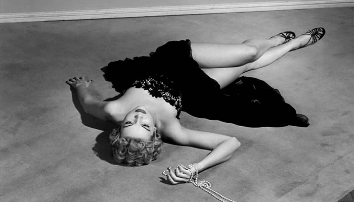A woman in a black dress is lying on the floor clutching a string of pearls.